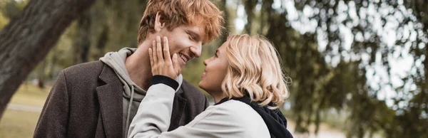 Homme rousse gai et femme blonde en manteau souriant pendant la date dans le parc, bannière — Photo de stock