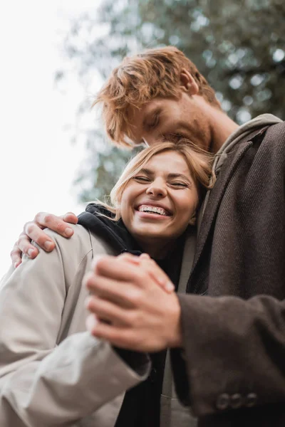 Low angle view of redhead man kissing head of happy blonde woman during date in park — Stock Photo