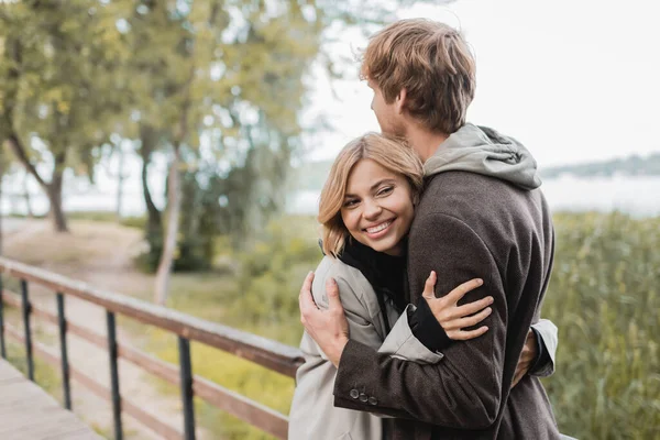Весела молода жінка посміхається і обіймає рудий чоловік під час побачення на мосту біля ставка — стокове фото