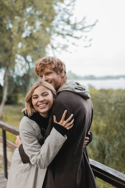 Gioiosa giovane donna sorridente e abbracciando rosso uomo durante la data sul ponte vicino stagno — Foto stock