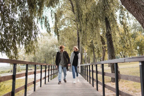 Full length of redhead man and blonde woman in coats smiling while holding hands and walking on bridge — Stock Photo