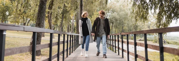 Longitud completa de pelirrojo hombre y mujer rubia en abrigos sonriendo mientras toma de la mano y caminar en el puente, bandera - foto de stock