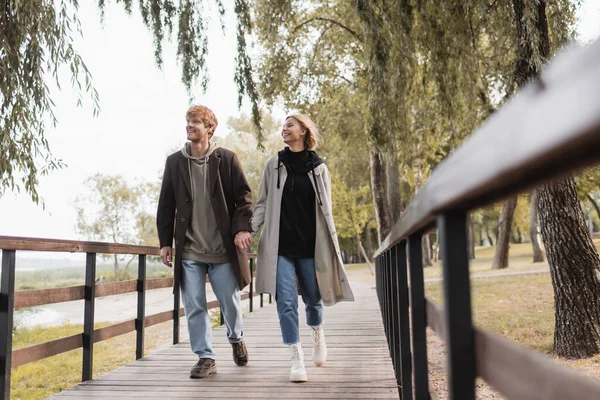 Longitud completa de feliz pareja en abrigos tomados de la mano y caminando en el puente - foto de stock