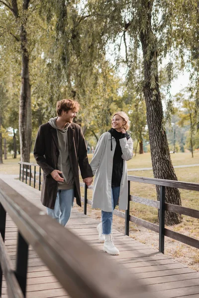 Full length of happy couple in coats holding hands and walking on bridge in park — Stock Photo