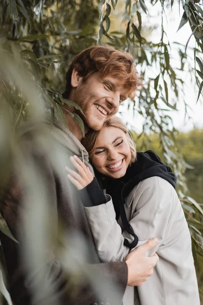 Feliz rubia mujer abrazando alegre joven hombre cerca verde hojas en borrosa primer plano - foto de stock