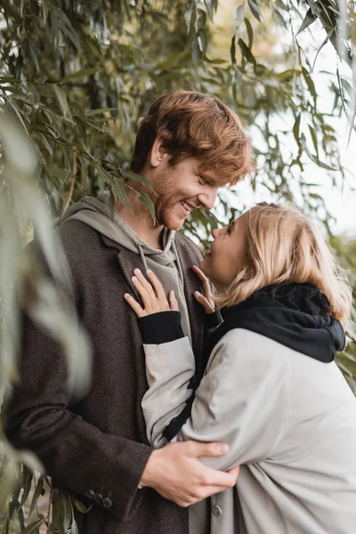 Heureuse femme blonde regardant joyeux jeune homme près de feuilles vertes sur le premier plan flou — Photo de stock