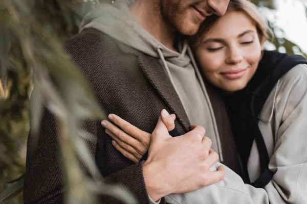 Heureux femme blonde étreignant avec joyeux jeune homme près de feuilles vertes sur le premier plan flou — Photo de stock