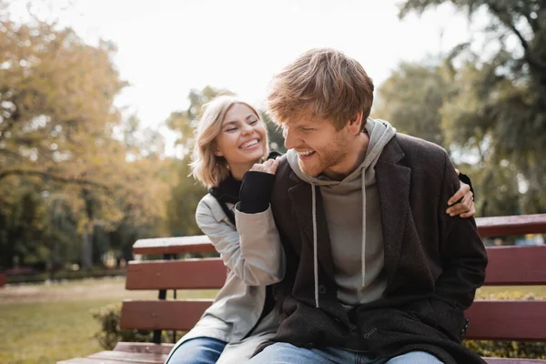 Happy and blonde woman hugging redhead boyfriend while sitting on bench in park — Stock Photo