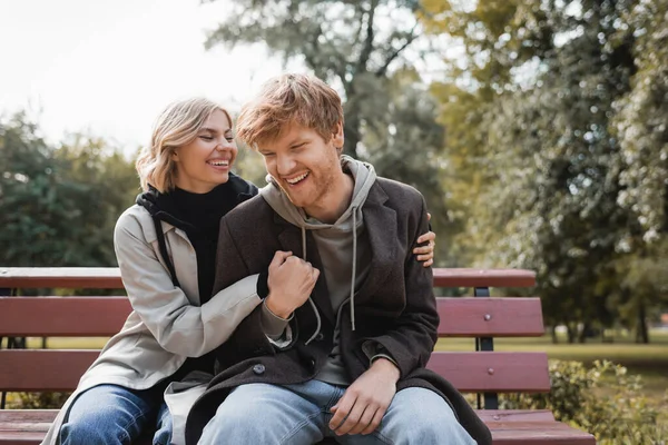 Cheerful and blonde woman embracing redhead boyfriend while sitting on bench in park — Stock Photo