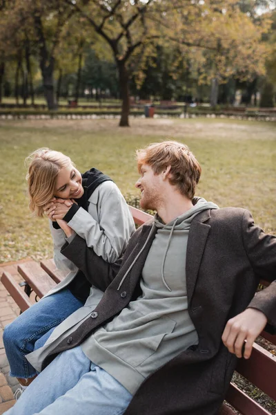 Mujer alegre y rubia abrazando la mano del novio pelirrojo mientras está sentado en el banco en el parque - foto de stock