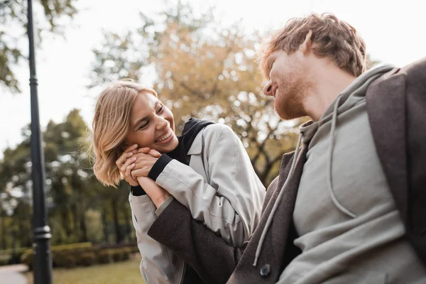Mulher alegre e loira segurando a mão do namorado ruivo enquanto sorri no parque — Fotografia de Stock