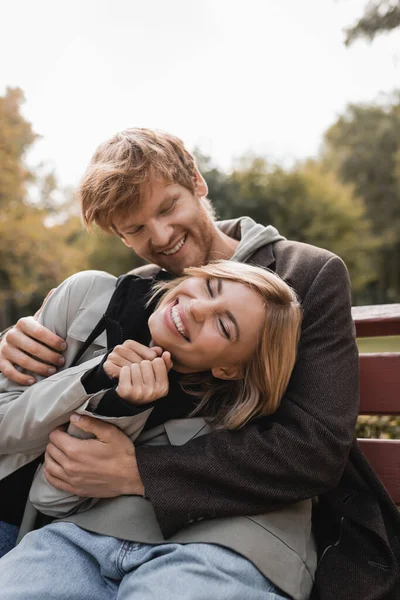 Rossa giovane uomo che abbraccia gioiosa donna bionda seduta sulla panchina nel parco autunnale — Foto stock