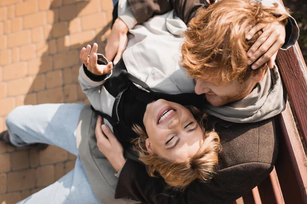 Vue de dessus de gai et rousse homme embrassant femme blonde heureuse tout en étant assis sur le banc dans le parc — Photo de stock