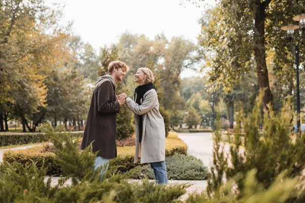 Side view of joyful and young couple in coats holding hands while standing around plants in park — Stock Photo