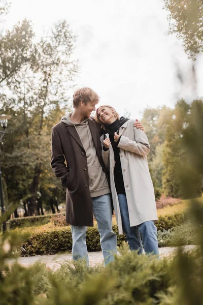 Happy and young man in coat hugging blonde girlfriend while standing around plants in park — Stock Photo