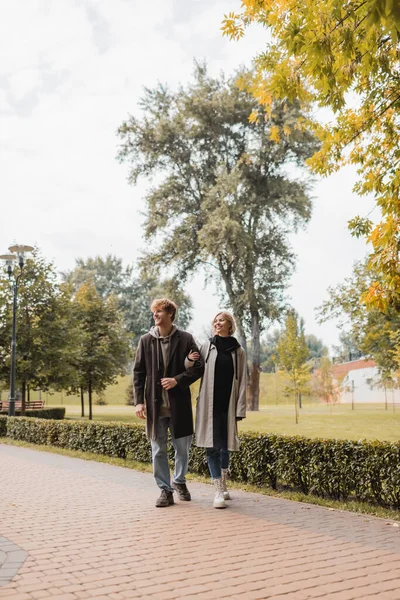 Full length of young and positive couple in coats walking together in autumnal park during date — Stock Photo