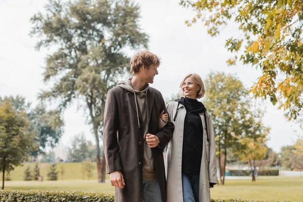 Jeune et positif couple en manteaux regarder l'autre tout en marchant dans le parc automnal pendant la date — Photo de stock