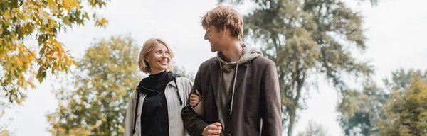 Positive couple in coats looking at each other while walking in autumnal park during date, banner — Stock Photo