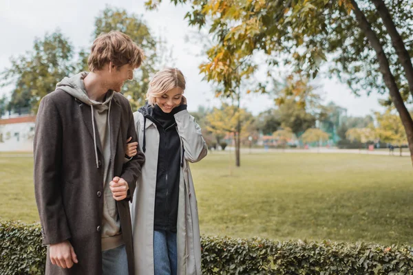 Young and positive man in coat walking with blonde girlfriend in autumnal park during date — Stock Photo