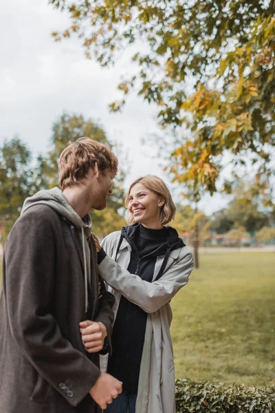 Pleased couple in coats looking at each other while walking in autumnal park during date — Stock Photo