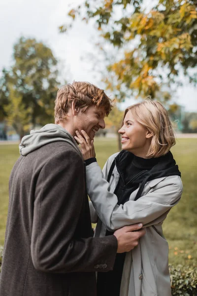 Joyful couple in coats looking at each other while embracing in autumnal park during date — Stock Photo