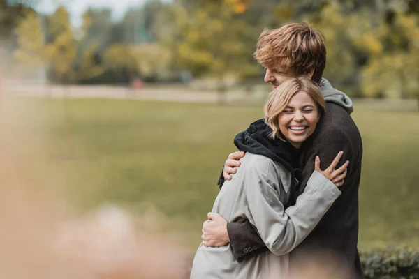 Cheerful young man and woman in autumnal coats embracing each other during date in park — Stock Photo