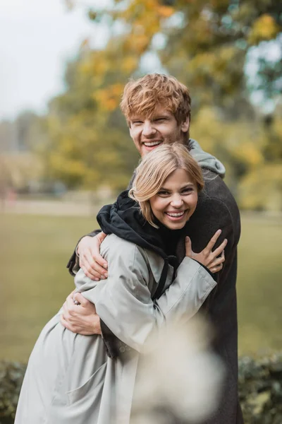 Fröhliche junge Mann und Frau in herbstlichen Mänteln umarmen einander und lächeln im Park — Stockfoto