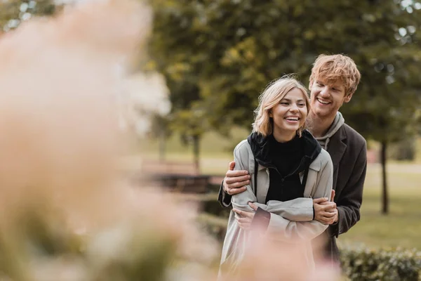 Positiver rothaariger Mann umarmt glückliche Frau, während er bei einem Date im Park zusammen lächelt — Stockfoto