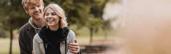 Positiver rothaariger Mann umarmt glückliche Frau, während er beim Date im Park zusammen lächelt, Banner — Stockfoto