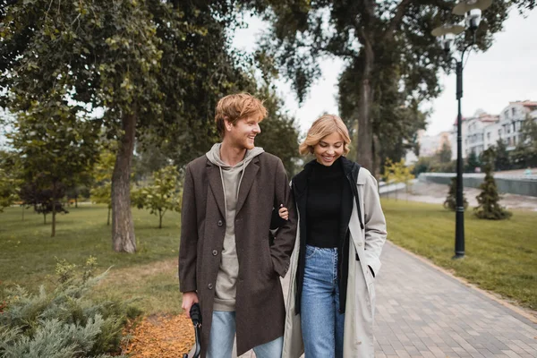 Hombre joven y positivo en abrigo caminando con novia rubia mientras sostiene el paraguas en el parque otoñal - foto de stock