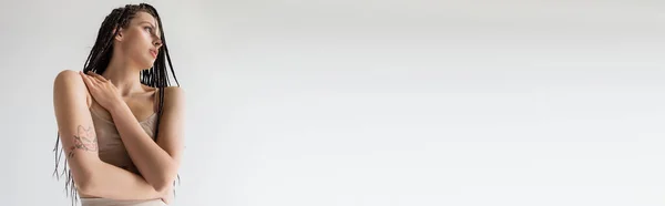 Low angle view of tattooed woman in sports bra looking away isolated on grey, banner — Stock Photo