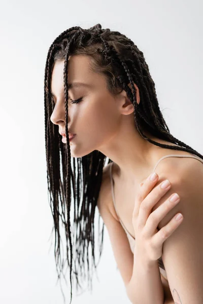 Joven morena con trenzas tocando hombro aislado en gris - foto de stock