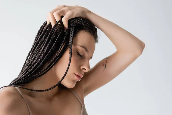 Портрет молодой татуированной женщины с косичками, касающимися волос, изолированных на сером — стоковое фото
