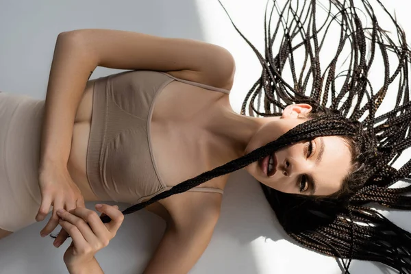 Visão superior da jovem mulher em sutiã esportivo tocando tranças no fundo cinza — Fotografia de Stock