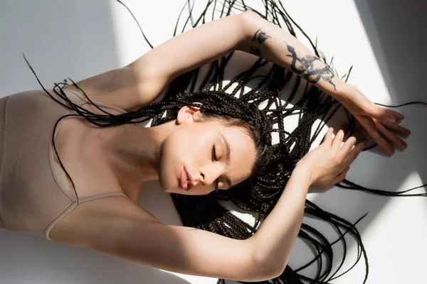 Вид зверху татуйованої жінки з косами, що лежать на сірому фоні зі світлом — стокове фото