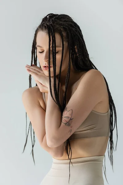 Young tattooed woman in sports bra holding hands near face isolated on grey — Stock Photo