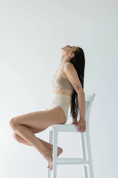 Side view of fit woman with braids sitting on chair isolated on grey — Stock Photo