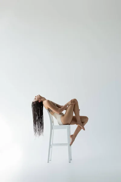 Side view of slim model in beige underwear sitting on chair on grey background — Stock Photo