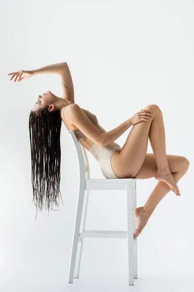 Vue latérale de la jeune femme avec des tresses assises sur une chaise sur fond gris — Photo de stock