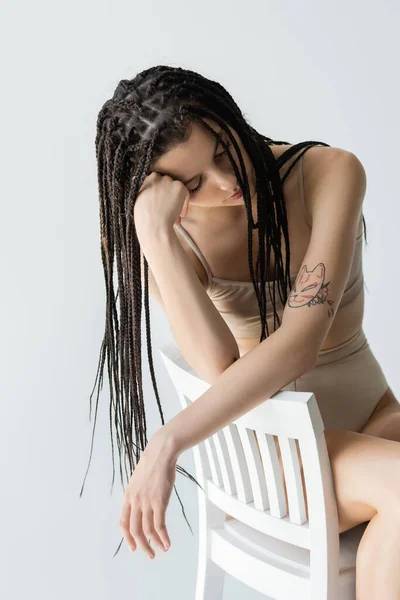 Femme tatouée avec des tresses assises sur une chaise blanche isolée sur gris — Photo de stock