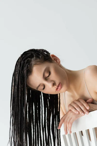 Jeune femme avec des tresses posant les yeux fermés près de chaise isolée sur gris — Photo de stock