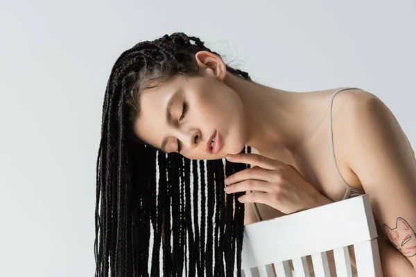 Tattooed woman with braids touching chin near chair isolated on grey — Stock Photo