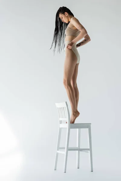 Seitenansicht einer fitten Frau in Unterwäsche, die auf einem Stuhl auf grauem Hintergrund steht — Stockfoto