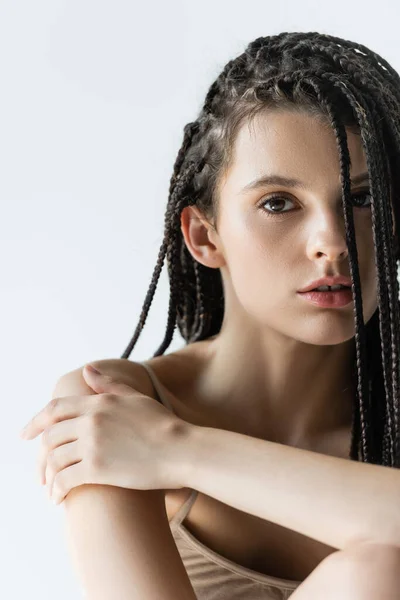 Retrato de mujer con trenzas tocando hombro y mirando a cámara aislada en gris - foto de stock