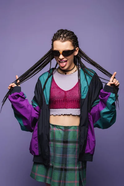 Mujer con estilo en ropa vintage tocando trenzas y sobresaliendo lengua aislada en púrpura - foto de stock