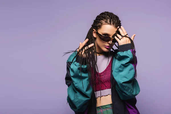 Mujer joven con trenzas peinado y ropa retro mirando a la cámara aislada en púrpura - foto de stock