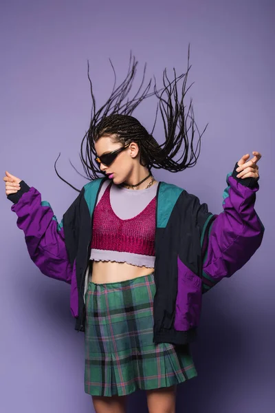 Стильная женщина в винтажной спортивной куртке и солнцезащитных очках позирует на фиолетовом фоне — стоковое фото