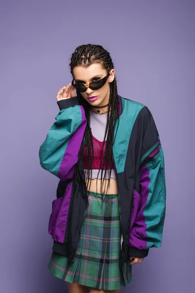 Jeune femme en veste de sport rétro tenant des lunettes de soleil sur fond violet — Photo de stock