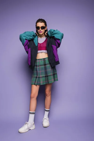 Modelo joven de moda en falda a cuadros y chaqueta deportiva de pie sobre fondo púrpura - foto de stock