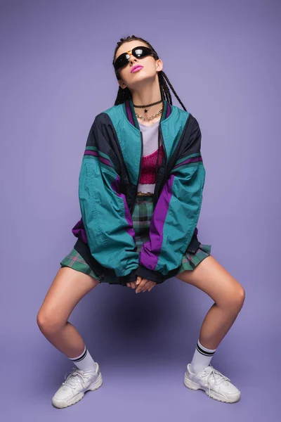 Mujer con estilo en gafas de sol y chaqueta de estilo noventa posando sobre fondo púrpura - foto de stock